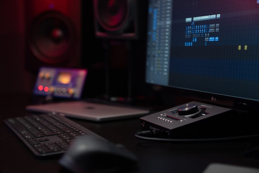 Photo of a computer monitor displaying audio software with a mouse, laptop, keyboard, and music controller in the foreground.